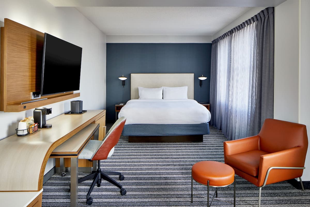 Habitación de hotel con cama, escritorio, silla, televisor y ventana con cortinas.