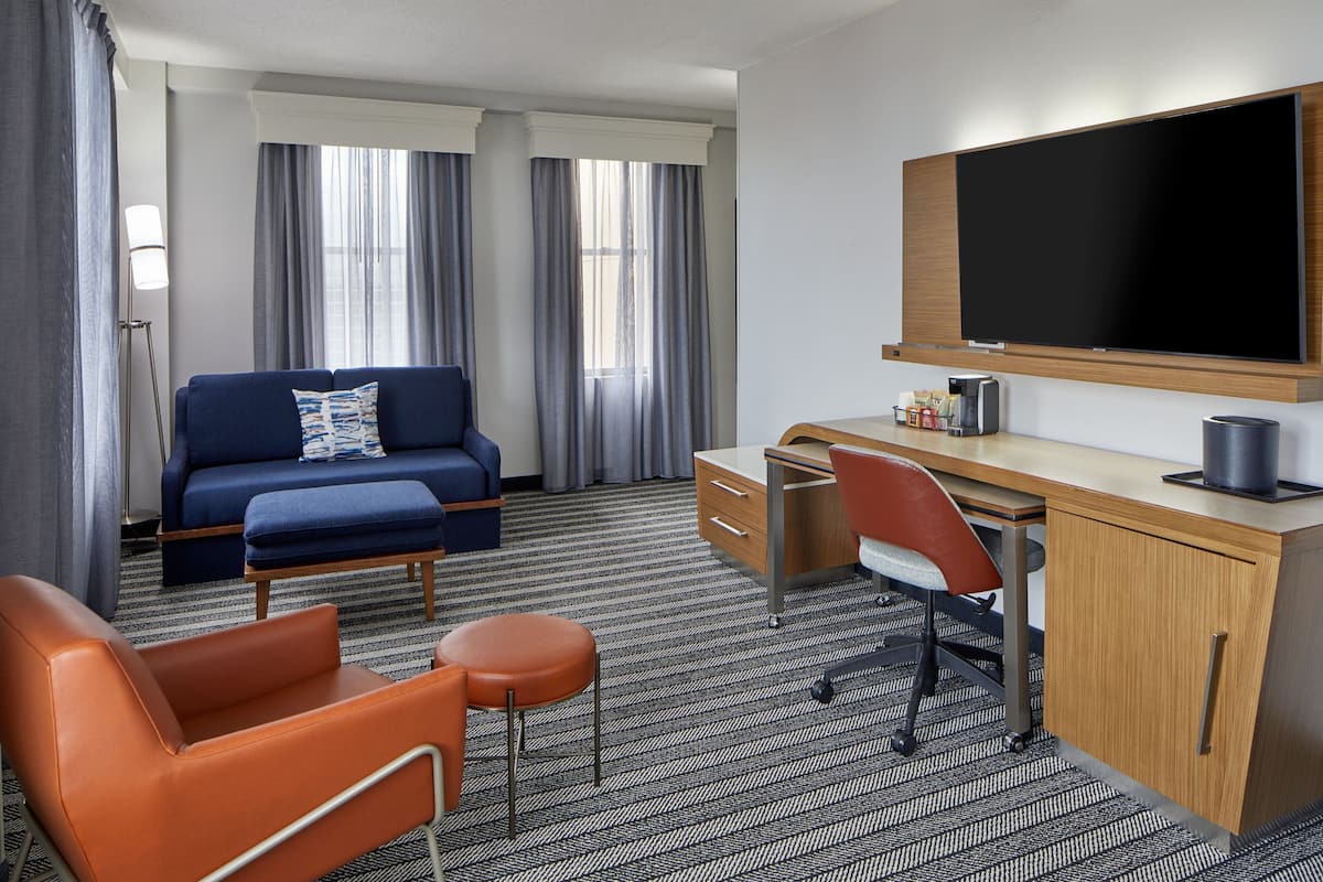 Habitación de hotel con sofá azul, sillón naranja, escritorio de madera y un televisor de pantalla plana grande.