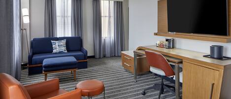 A hotel room with a blue sofa, orange armchair, wooden desk, and a large flat-screen TV.
