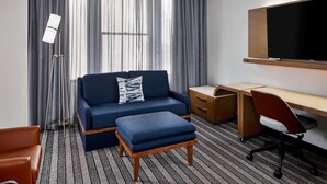 A hotel room with a sofa, ottoman, desk, chair, and a TV on the wall. - Courtyard By Marriott Nashville Downtown (Nashville)