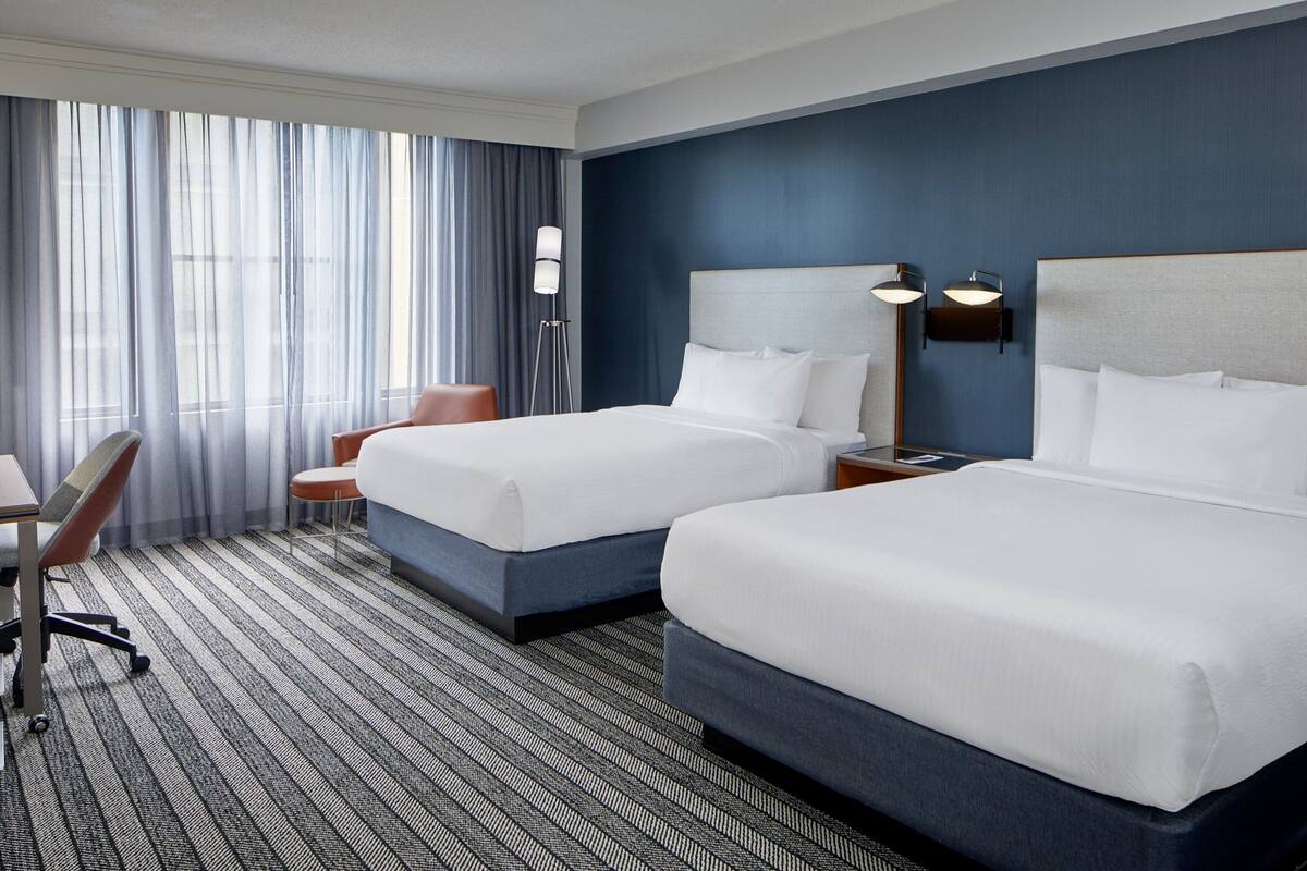 Habitación de hotel con dos camas, un escritorio, una silla y un ventanal con cortinas.