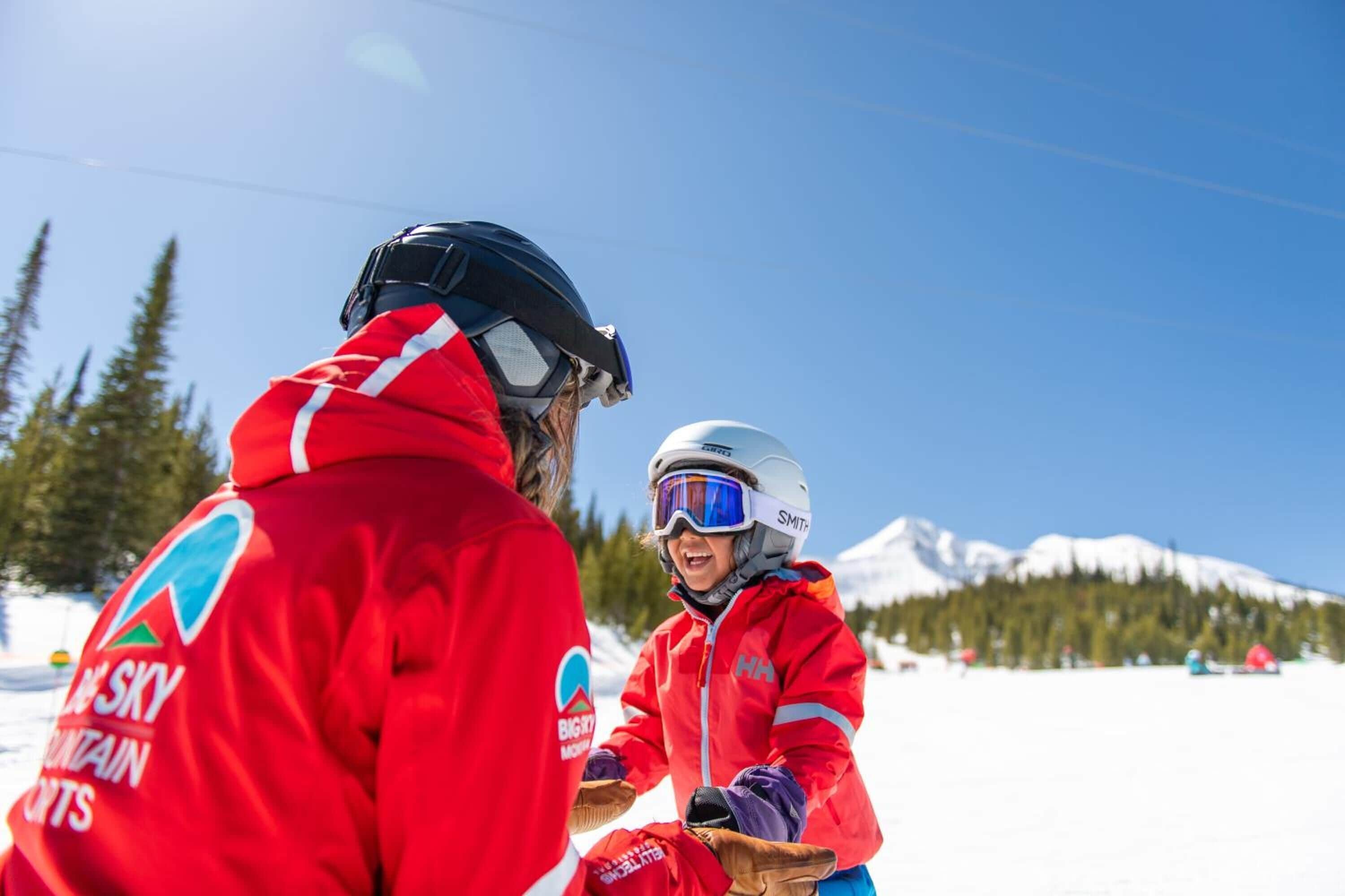 Two people in red jackets with logos, one adult and one child, in a snowy outdoor setting with mountains in the background.