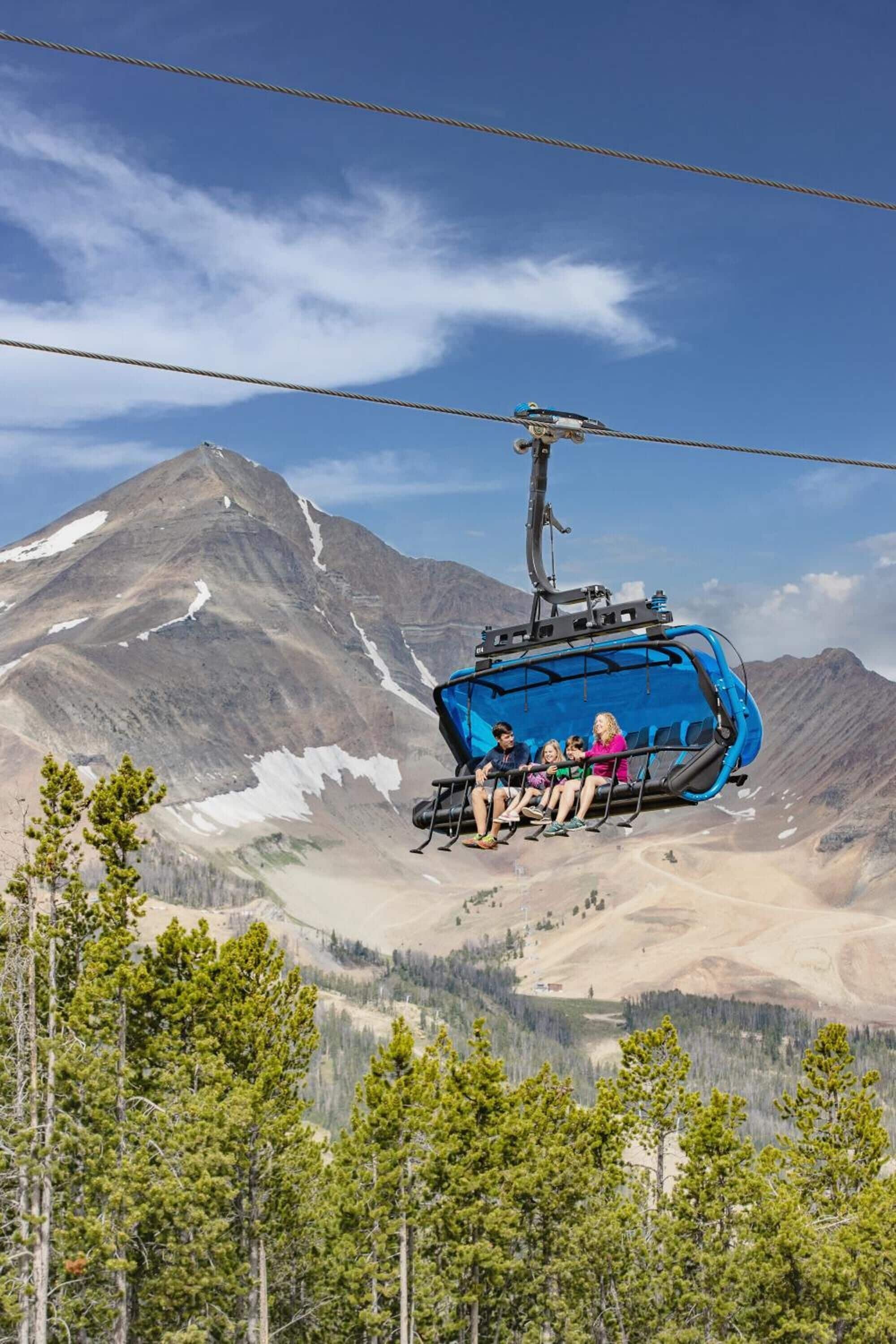 Eine blaue Seilbahn mit Fahrgästen, eine gebirgige Landschaft und spärliche Bäume im Vordergrund.