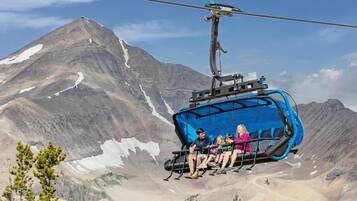 Eine blaue Seilbahn mit Fahrgästen, eine gebirgige Landschaft und spärliche Bäume im Vordergrund.