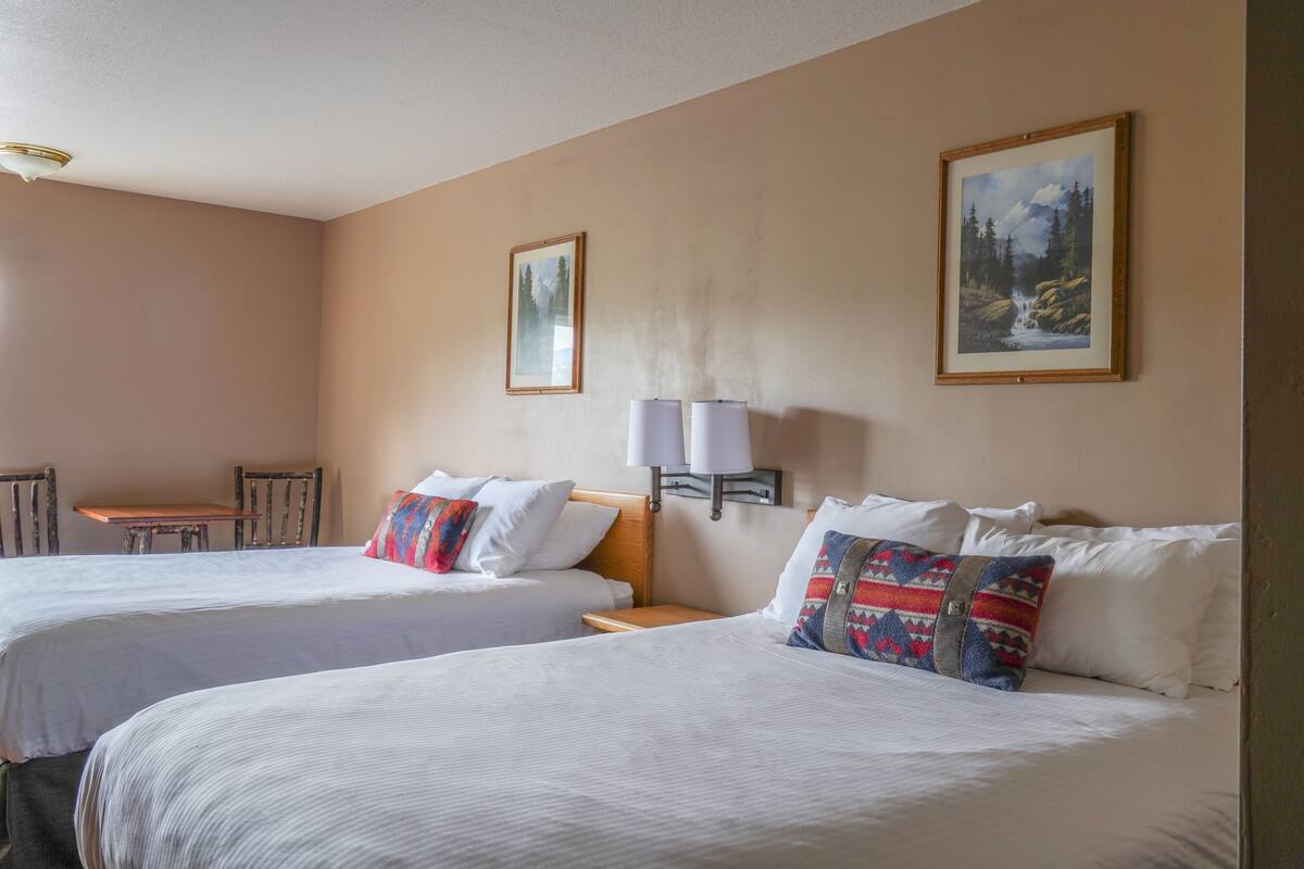 A hotel room with two beds, a small table, and two chairs. There are two wall-mounted lamps and two framed pictures on the wall.