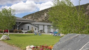 A building with a grey facade, a covered parking area, and a grassy lawn with a rock garden. - Whitewater Inn (Gallatin Gateway)