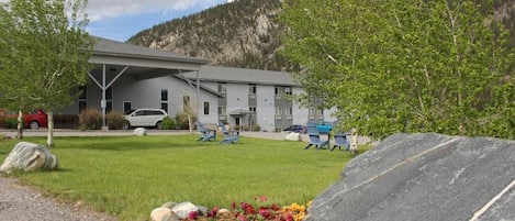 A building with a grey facade, a covered parking area, and a grassy lawn with a rock garden.