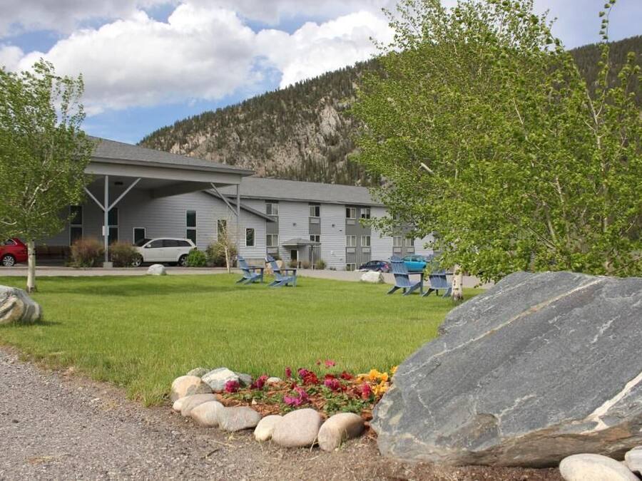 A building with a grey facade, a covered parking area, and a grassy lawn with a rock garden.