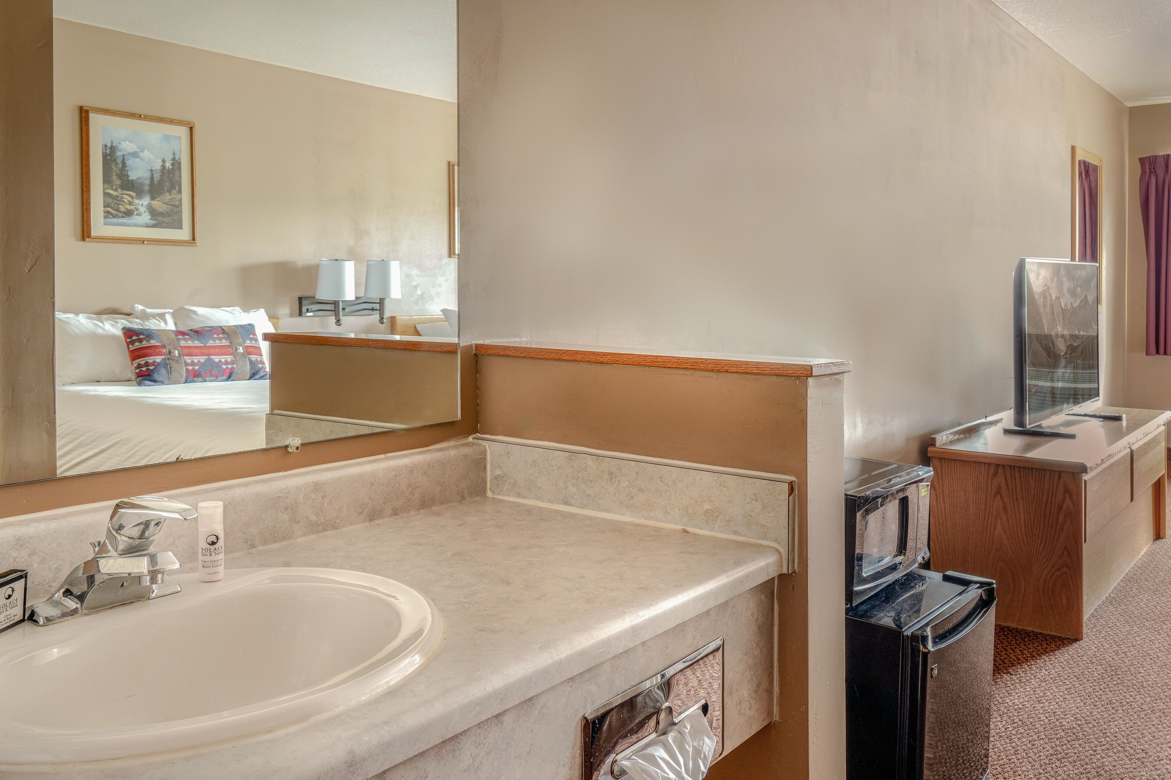 A hotel bathroom with a large sink, mirror, and a small television on a wooden cabinet.