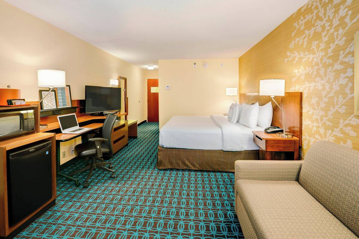 A hotel room with a bed, a desk with a laptop, a television, a desk lamp, a chair, and a small refrigerator.