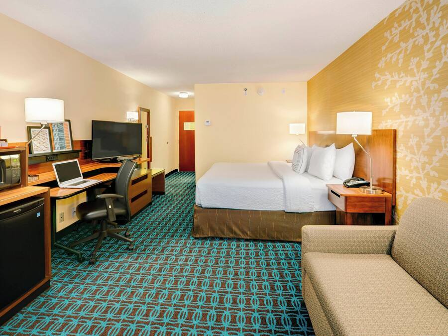 A hotel room with a bed, a desk with a laptop, a television, a desk lamp, a chair, and a small refrigerator.
