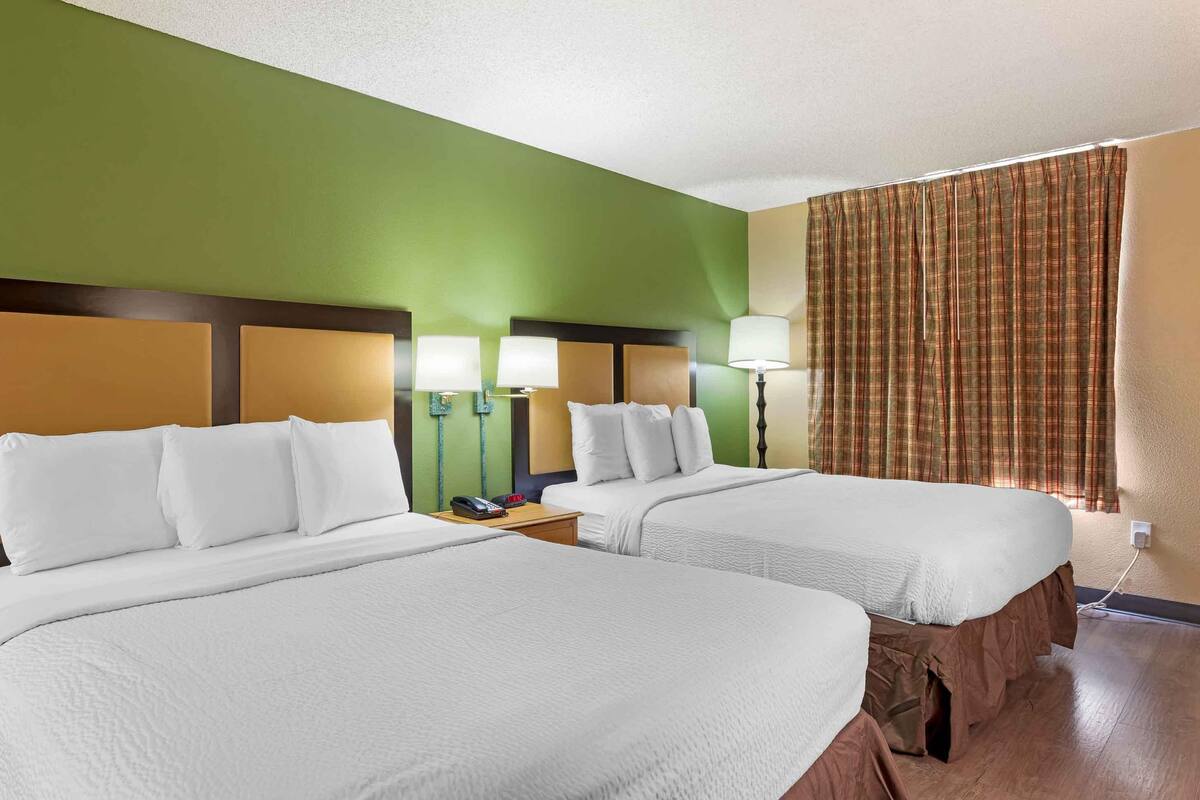 A hotel room with two beds, green walls, a brown headboard, a nightstand with a lamp, and a plaid curtain.