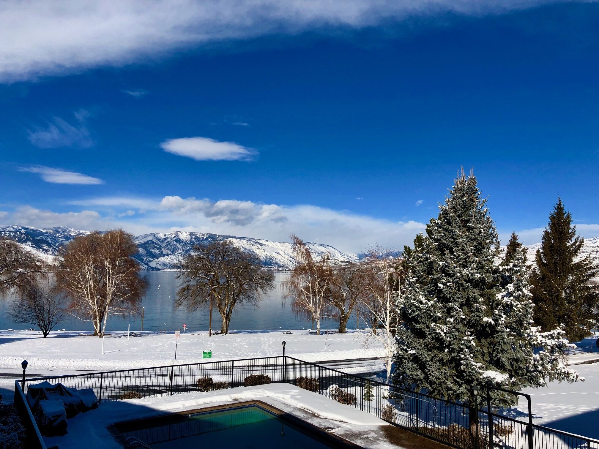 A snow-covered landscape with a clear blue sky, a frozen lake, and snow-covered mountains in the background.