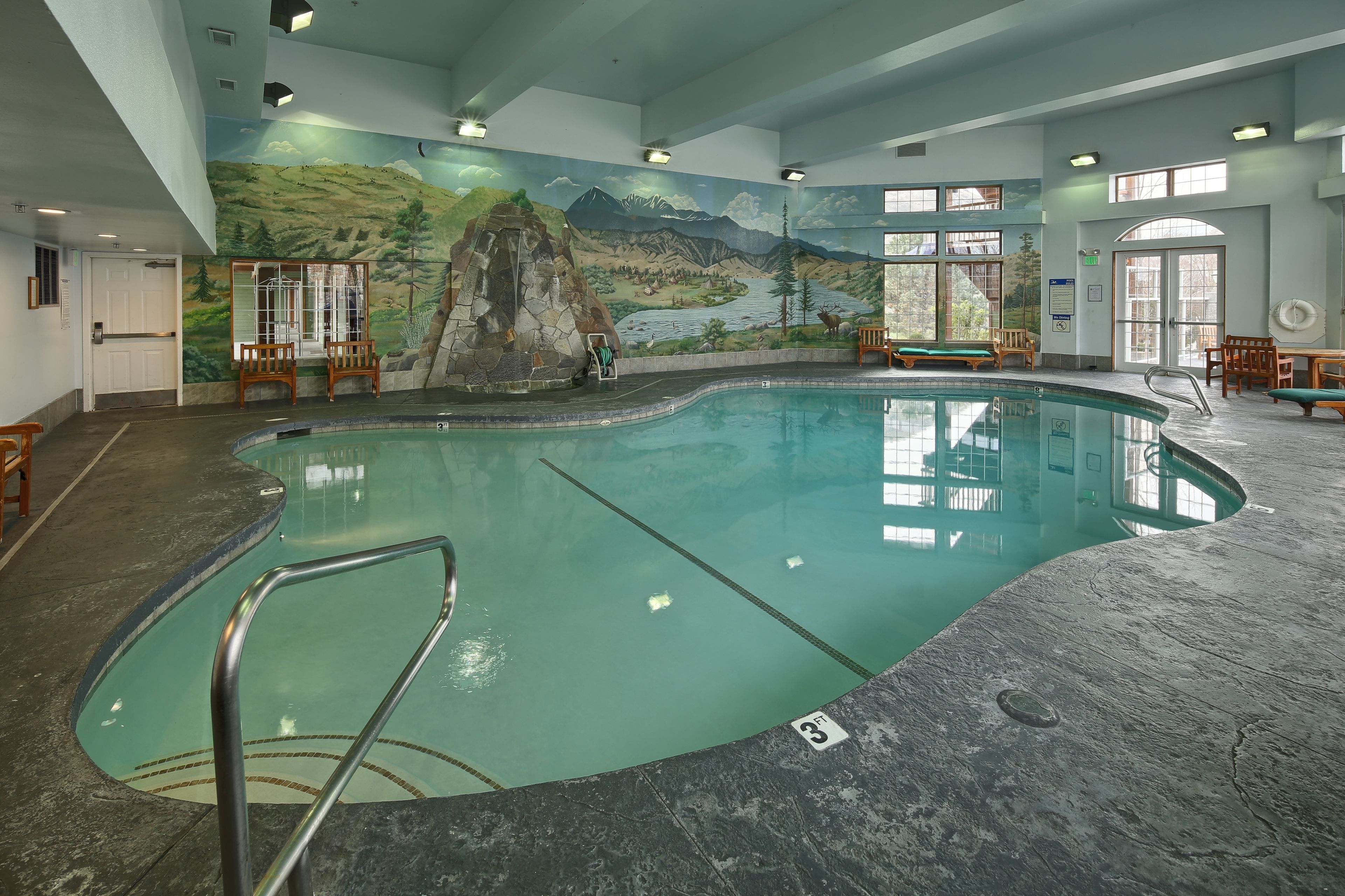 An indoor swimming pool with a mural of a mountainous landscape, wooden seating around the pool, and a clear view of the pool's edge.