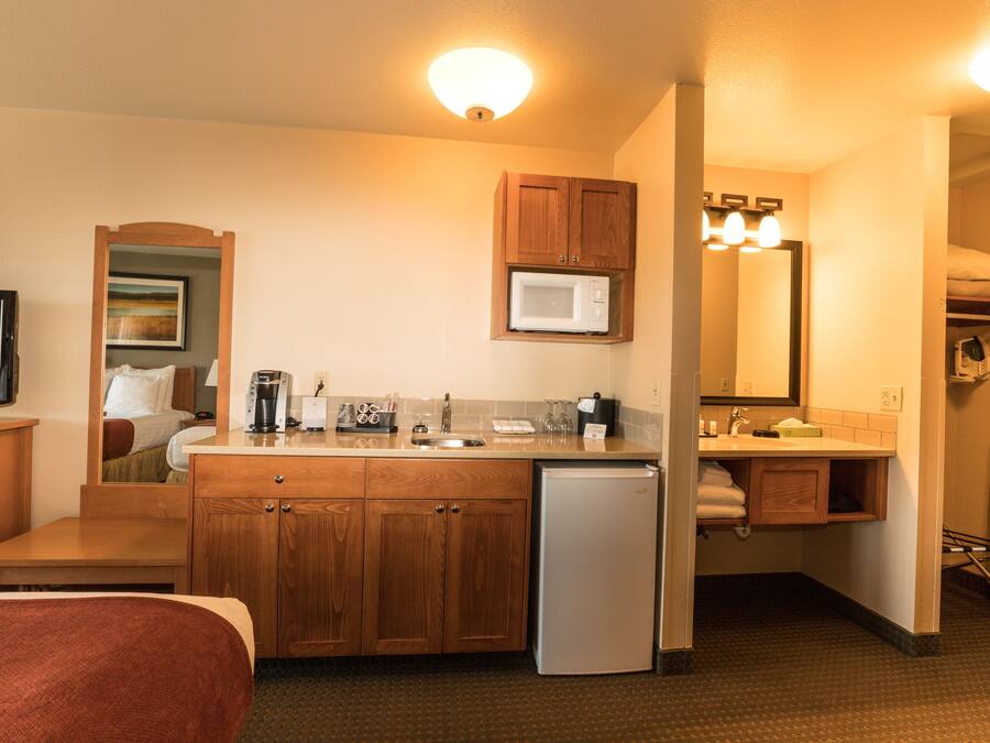 Habitación de hotel compacta con kitchenette, cama, televisión y baño con espejo y lavamanos.