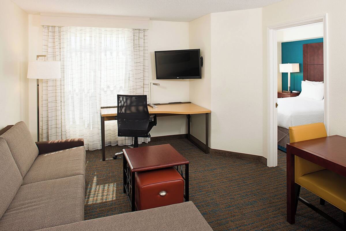A hotel room with a sofa, a desk with a chair, a television, and a bed with a nightstand.