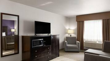 Habitación de hotel con televisión de pantalla plana sobre un mueble de madera, un sillón gris, un sofá, un espejo grande y una ventana con cortinas.