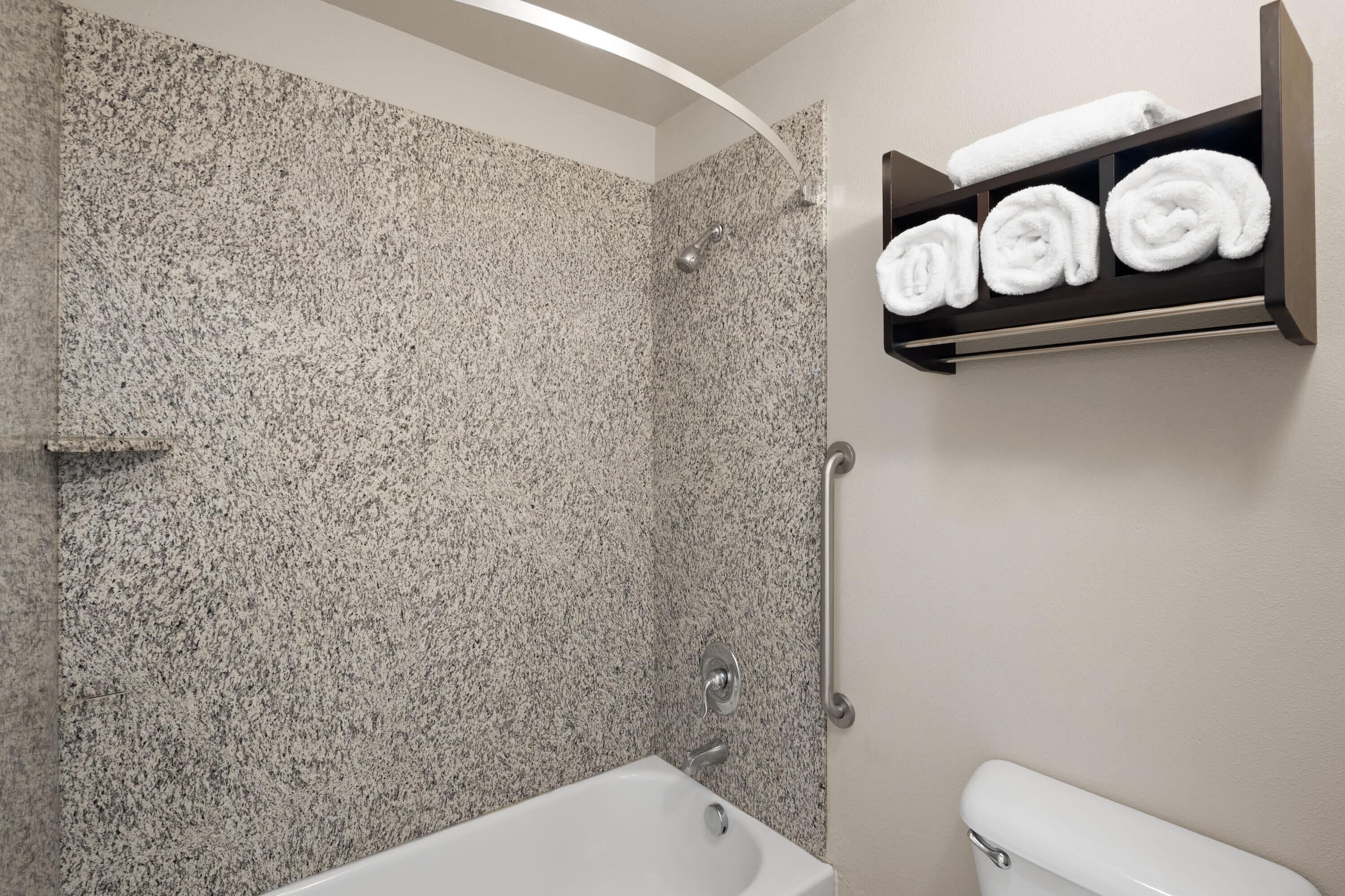 A bathroom with a granite shower wall, a white bathtub, and a towel rack with neatly rolled towels.