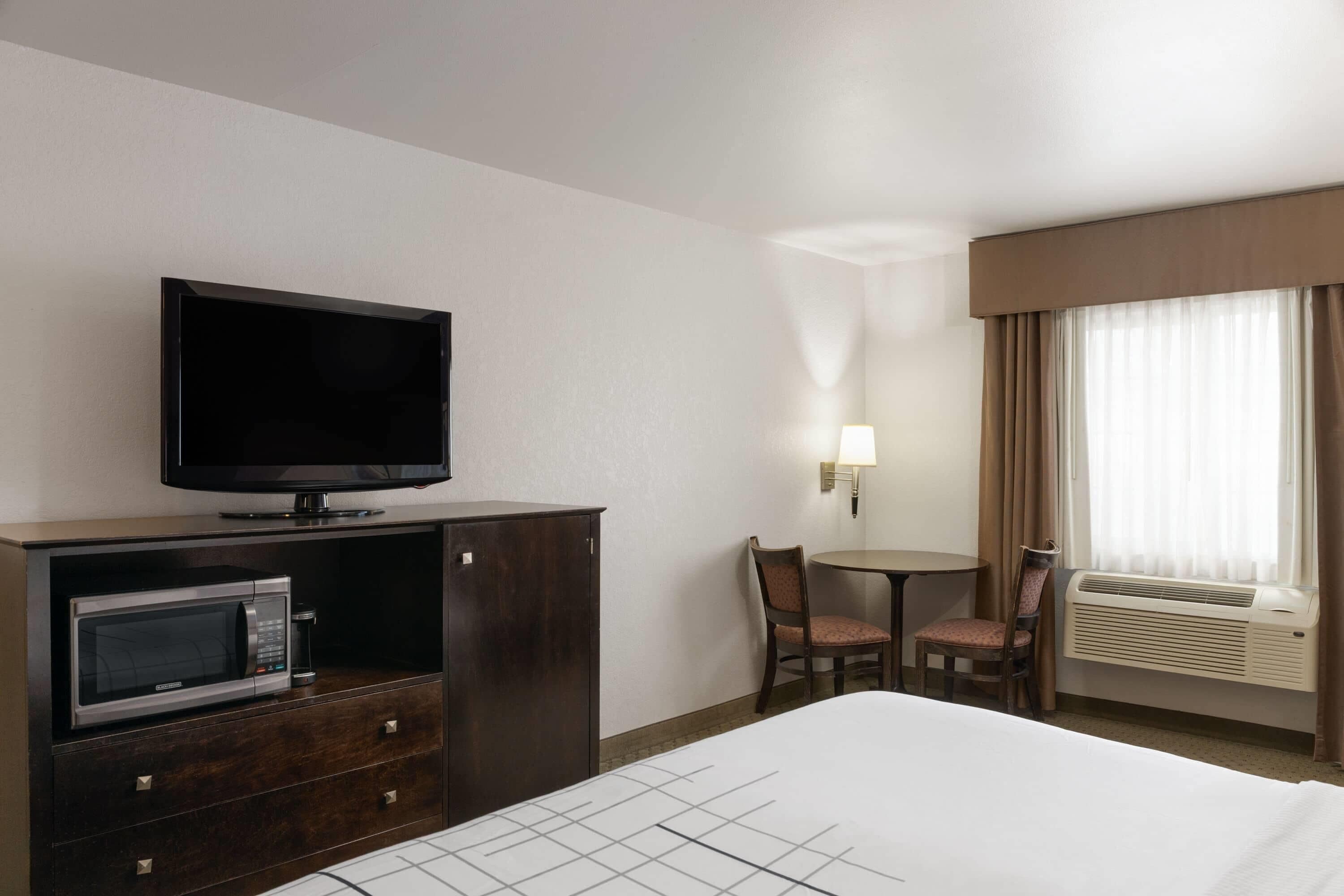 Habitación de hotel con cama, televisor en mueble bajo, microondas, mesa con sillas y ventana con cortinas.