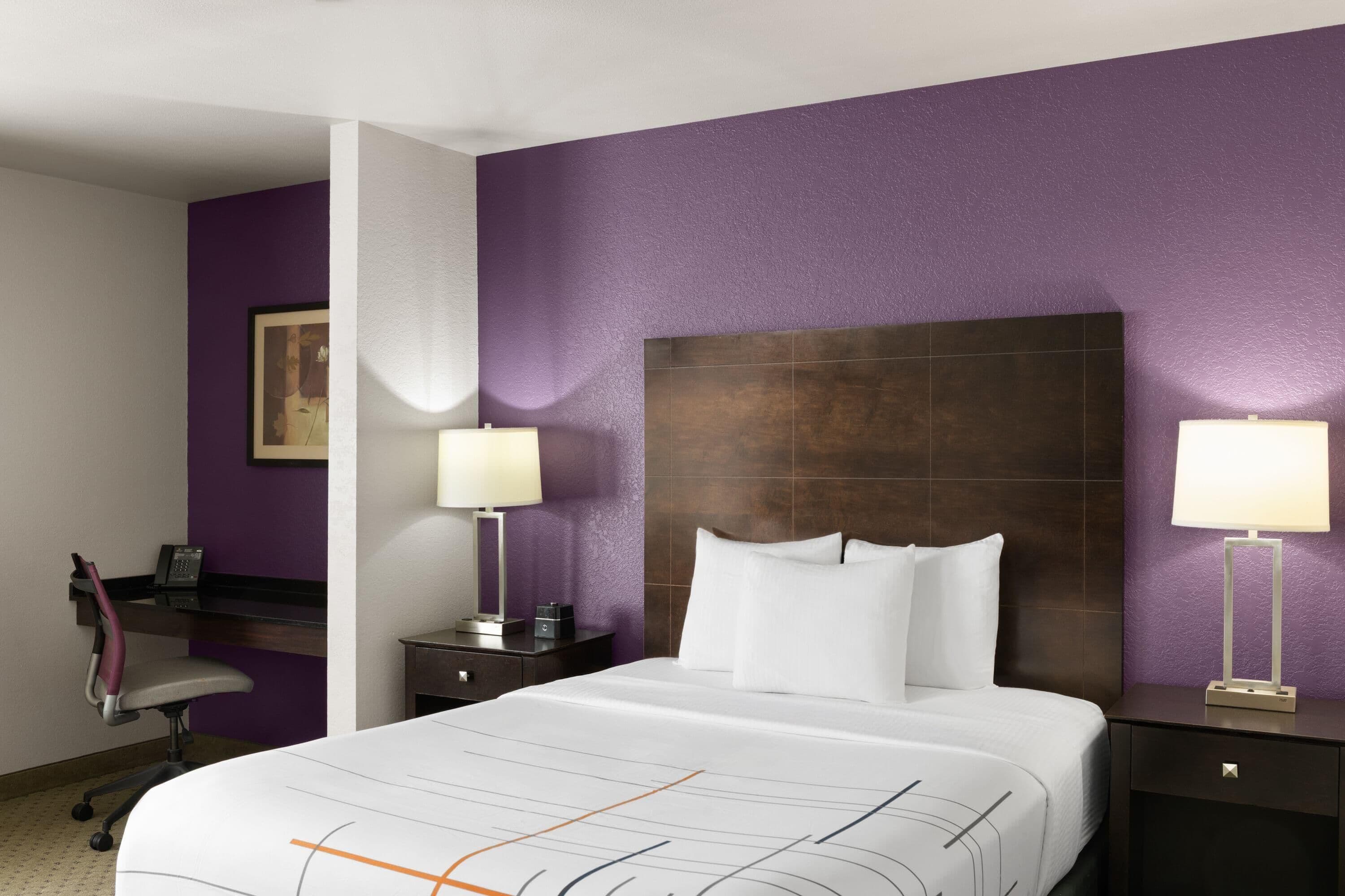 Una camera d'albergo con un letto grande, due comodini con lampade, una scrivania con sedia e pareti viola.