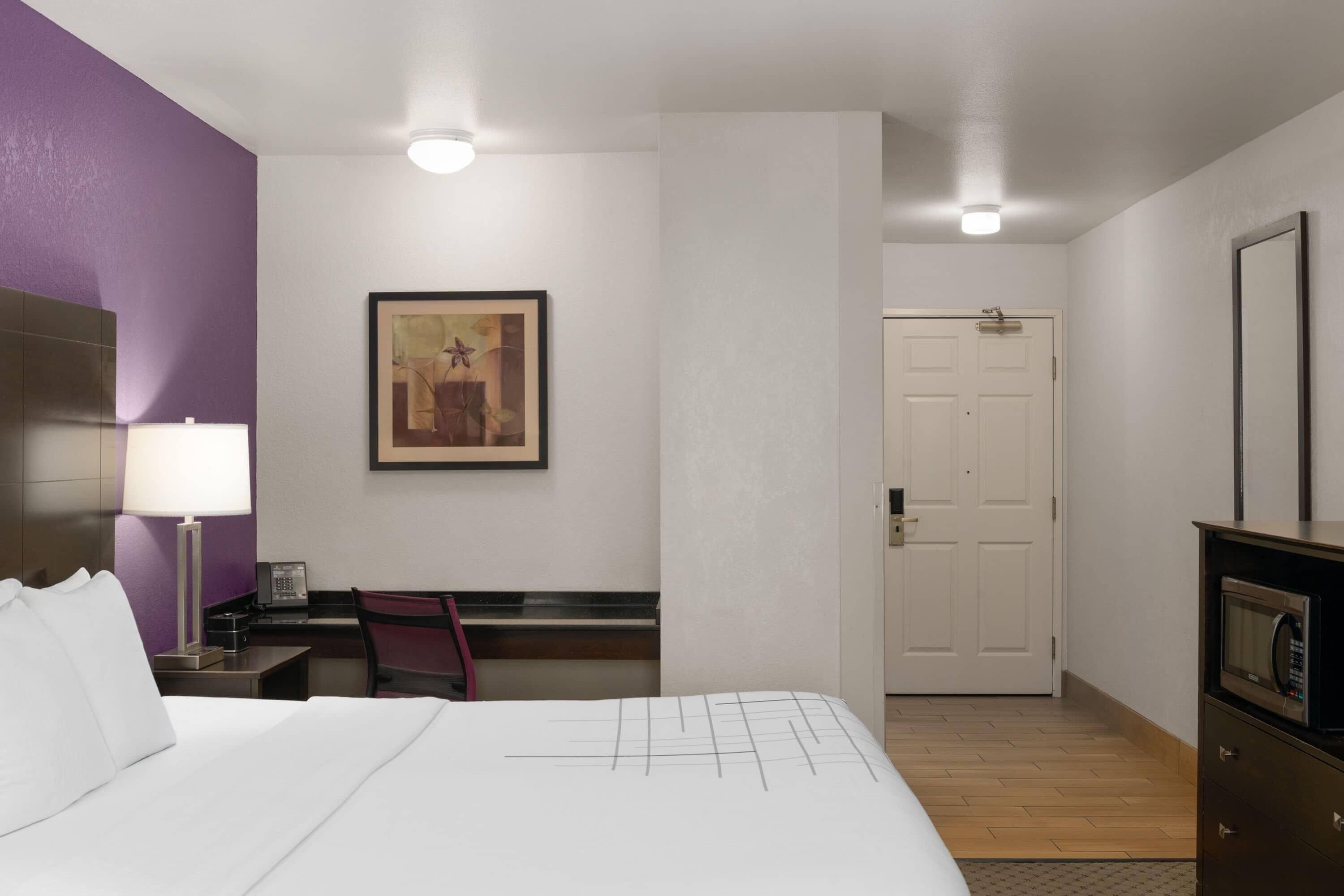 Una habitación de hotel con cama, escritorio, silla, lámpara de escritorio, microondas, puerta y un cuadro en la pared.