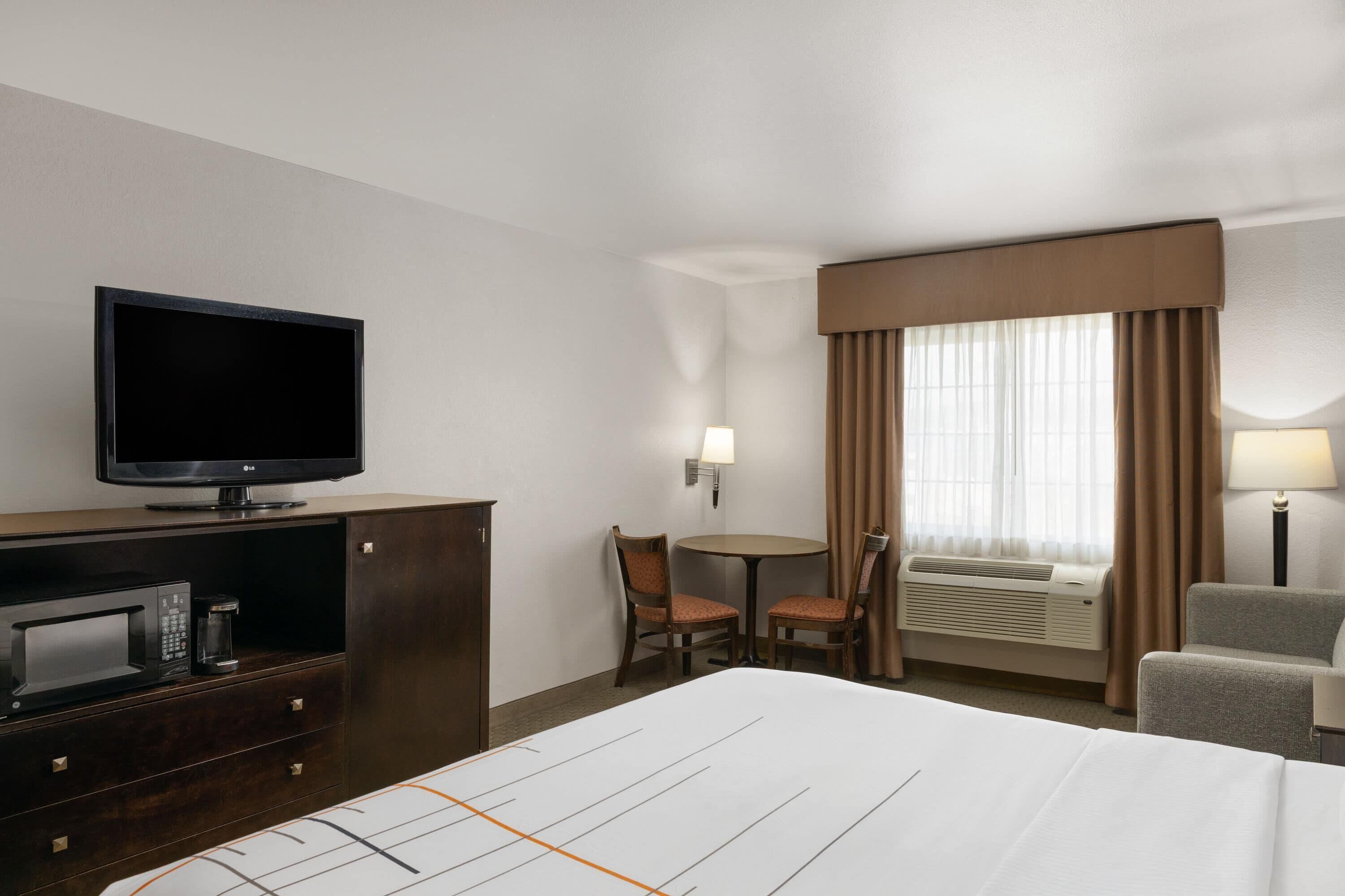 A hotel room with a bed, a TV, a microwave, a small table with chairs, and an air conditioning unit.