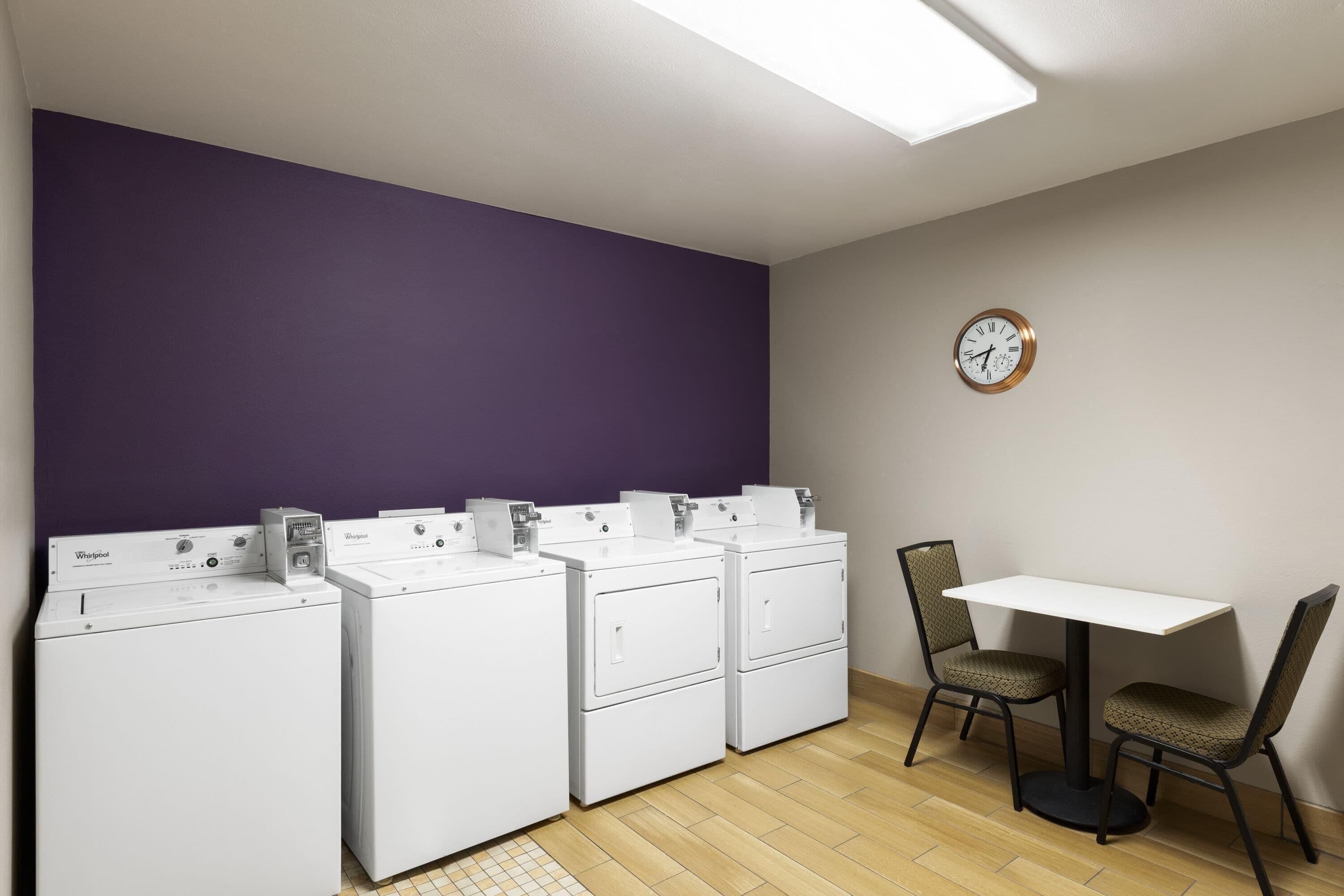 Une buanderie équipée de trois machines à laver et de trois sèche-linge superposés, une table avec deux chaises et une horloge au mur.