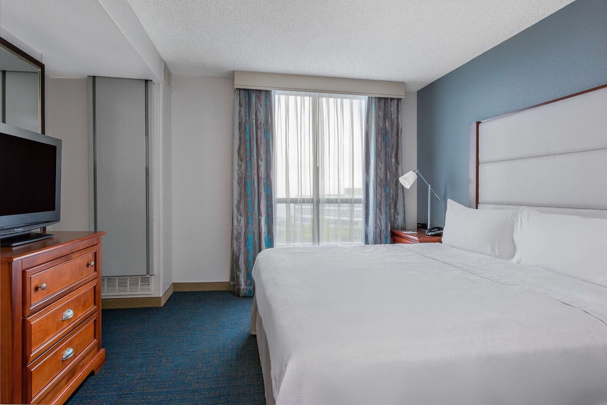 A hotel room with a bed, a TV on a dresser, a lamp, and a window with curtains.