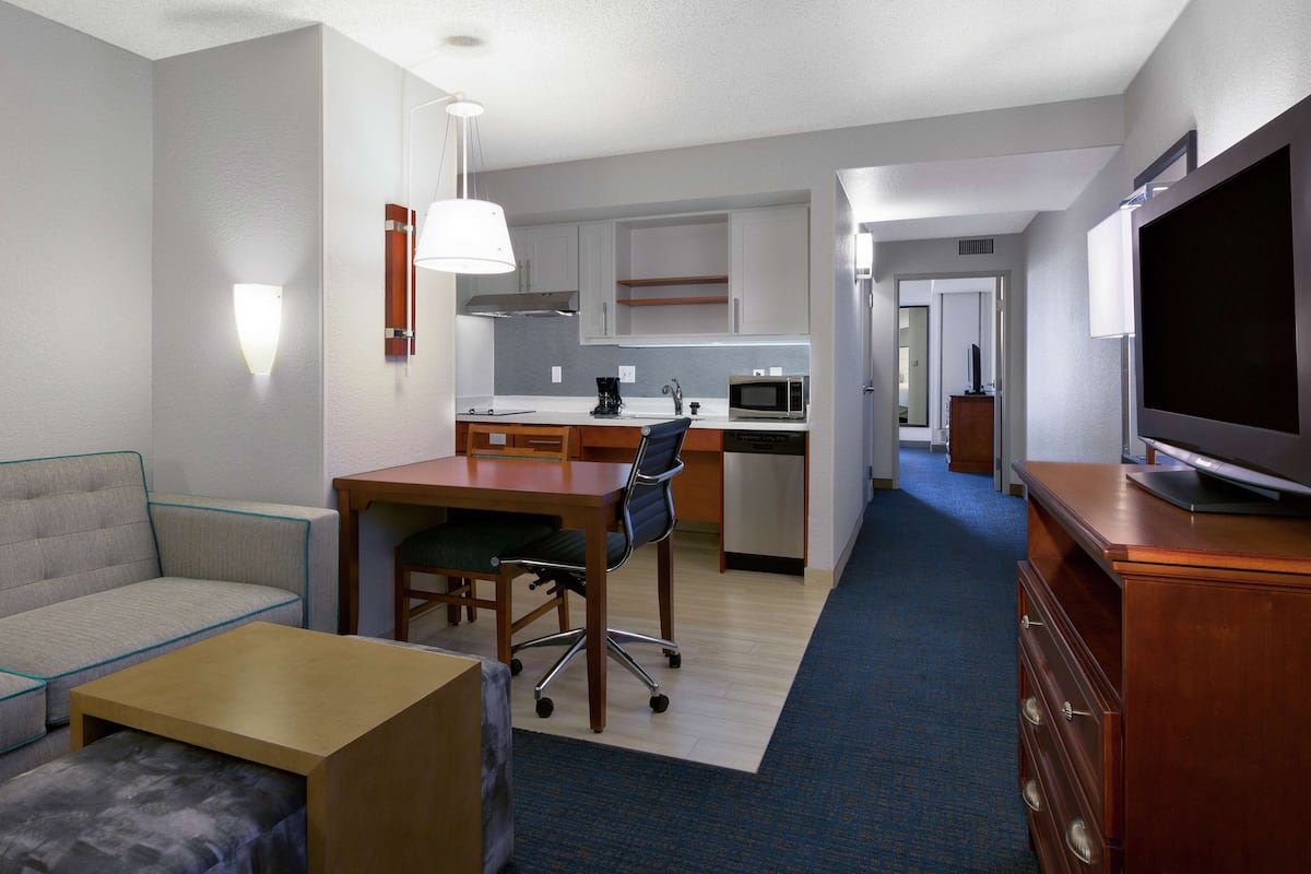 A hotel room with a sofa, a small table, a kitchenette, a TV, and a hallway leading to another room.