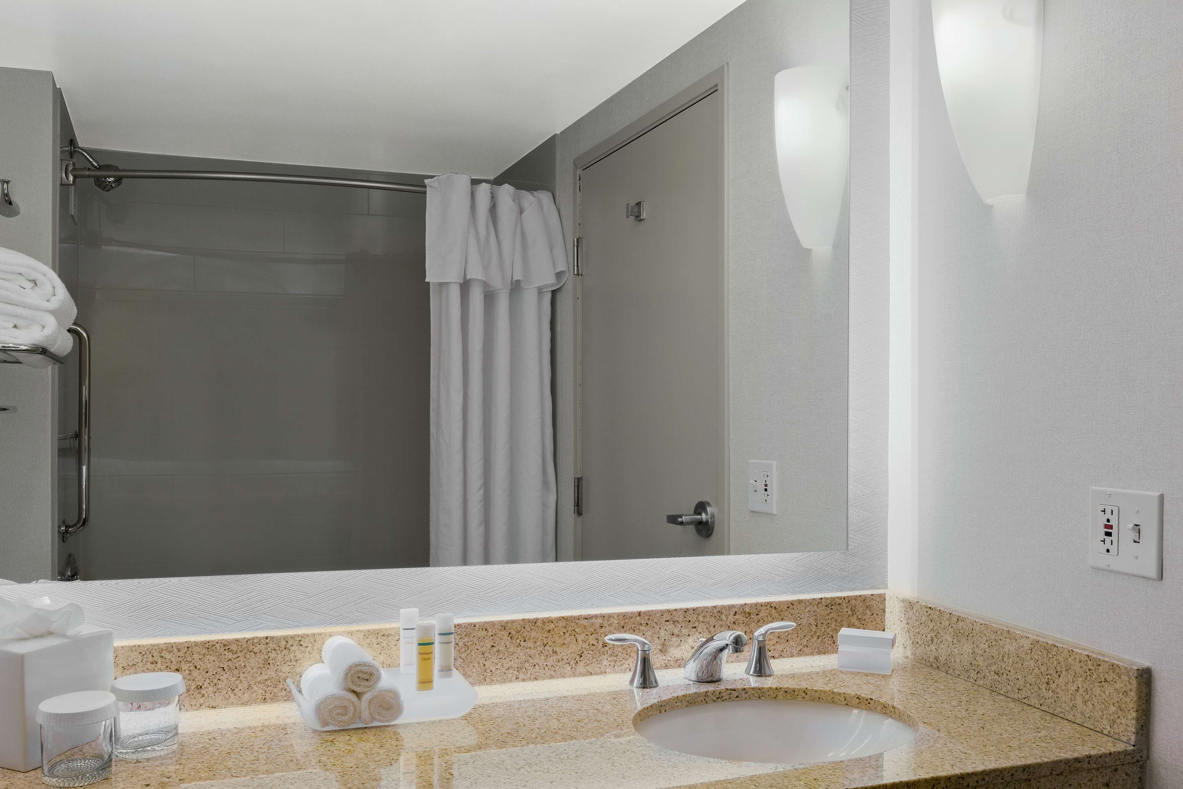 Een hotelbadkamer met een grote spiegel, een wastafel met een granieten aanrechtblad, een douche en keurig opgevouwen handdoeken.