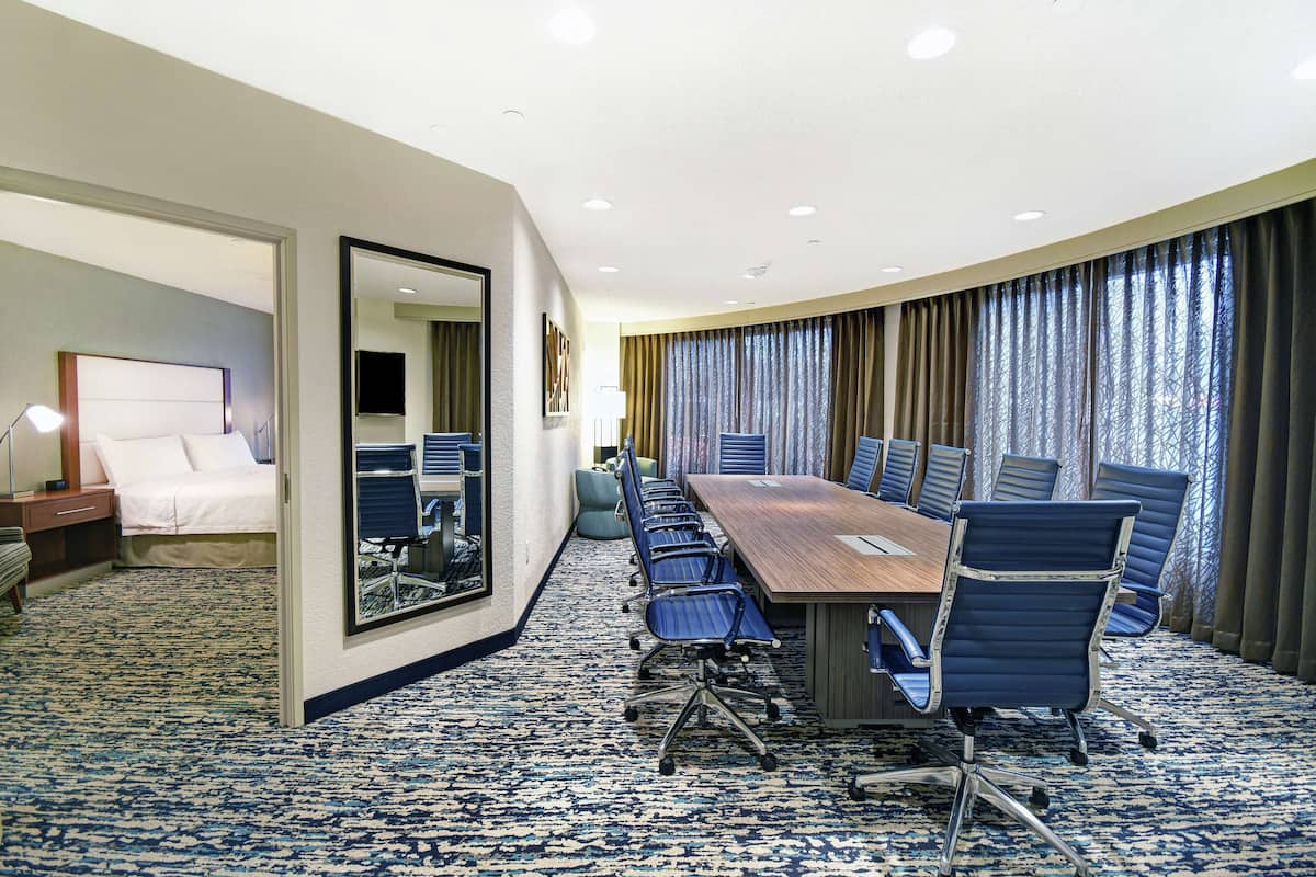 A hotel conference room with a long table, blue chairs, a bed, and a large mirror.