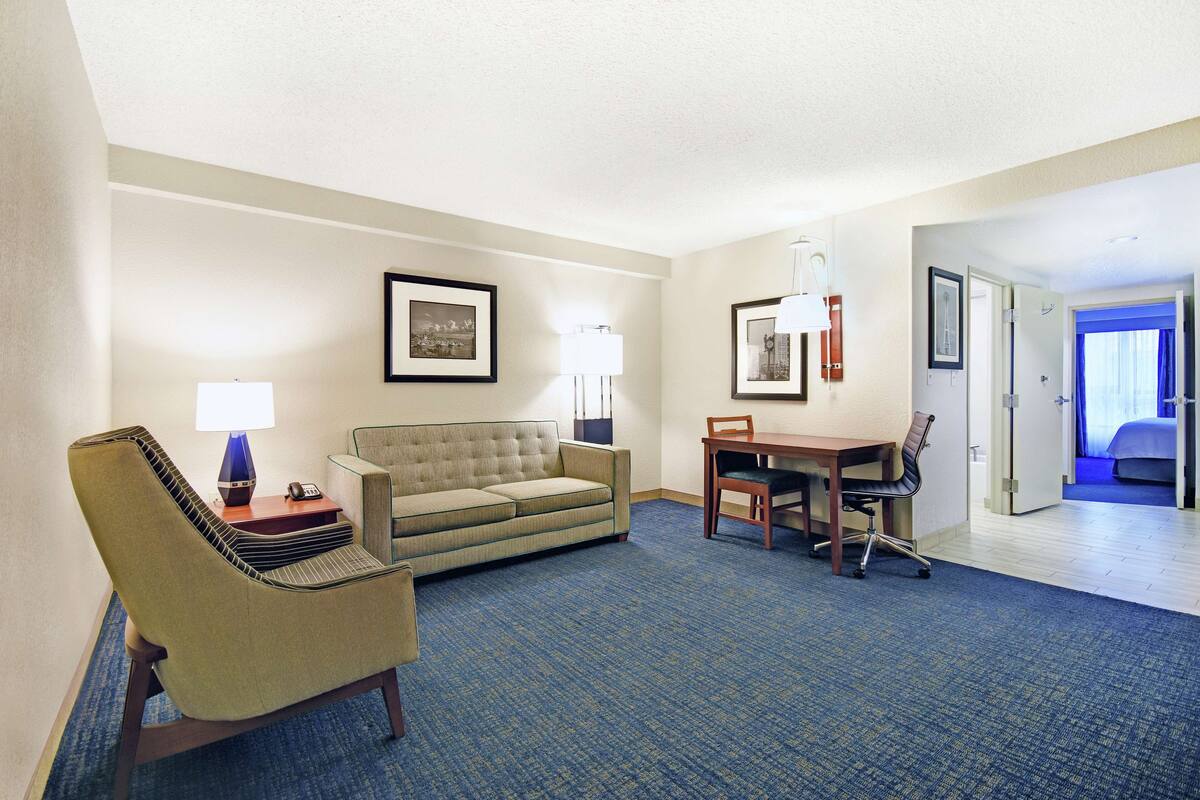 A hotel room with a sofa, armchair, dining table, and chairs. There are framed pictures on the walls and a lamp on the side table.