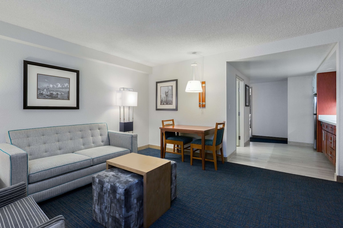 A hotel room with a grey sofa, a wooden dining table with chairs, a small coffee table, and framed pictures on the wall.