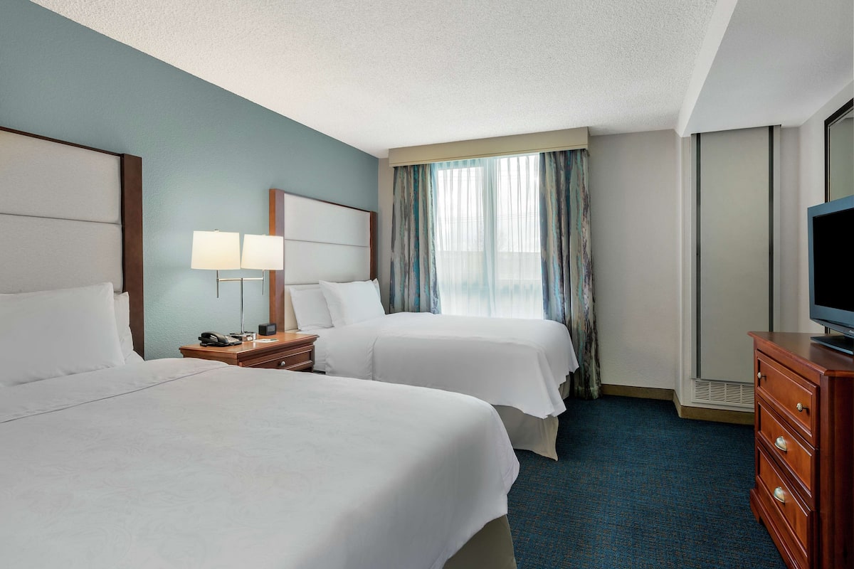 A hotel room with two beds, a nightstand with a lamp, a dresser with a TV, and a large window with curtains.