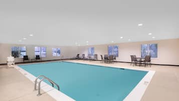 Une piscine intérieure à l’eau d’un bleu cristallin, entourée de chaises longues et de tables, et dotée de grandes fenêtres offrant une vue sur l’extérieur.