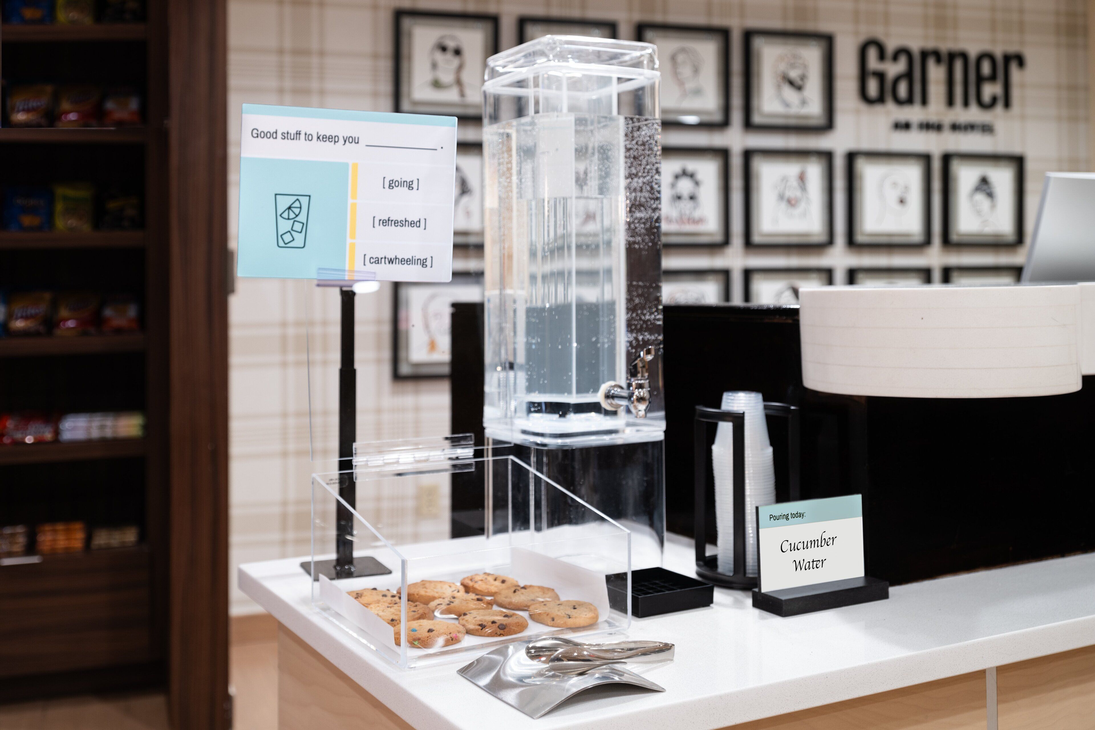 A coffee station with a water dispenser, a sign indicating cucumber water, and a display of cookies.