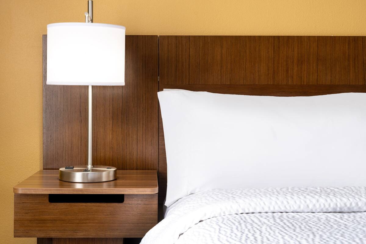A neatly made bed with white linens, a wooden nightstand with a lamp, and a wooden headboard.