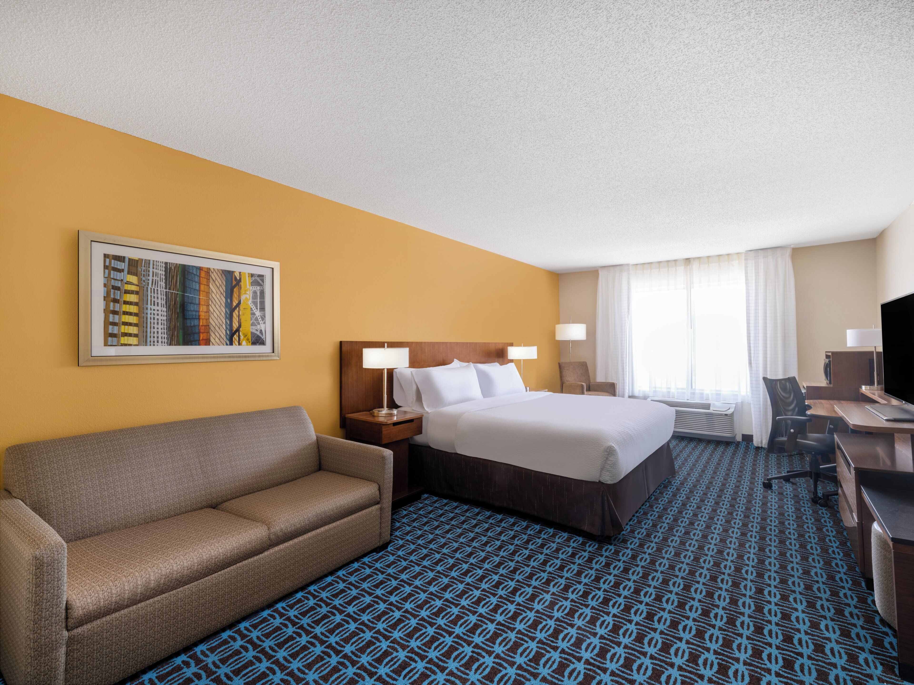 A hotel room with a bed, a sofa, a desk, a TV, and a painting on the wall.