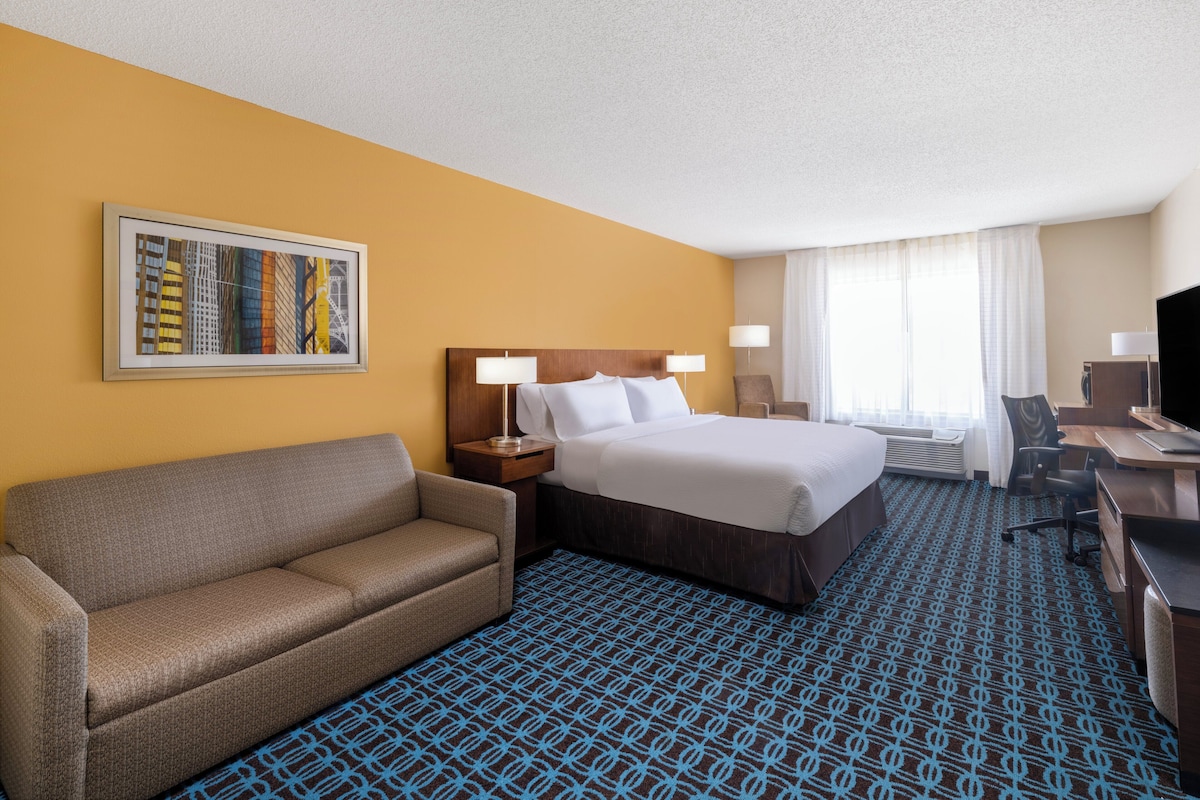 A hotel room with a bed, a sofa, a desk, a TV, and a painting on the wall.