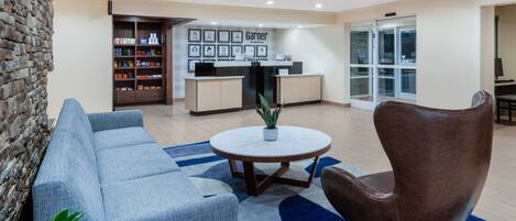 A modern hotel lobby with a reception desk, a blue sofa, a brown chair, and a round table with a plant.