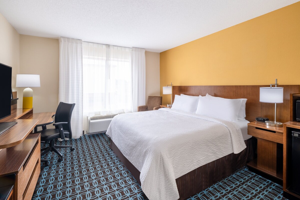 A hotel room with a large bed, a desk with a computer, a chair, a TV, and a window with curtains.