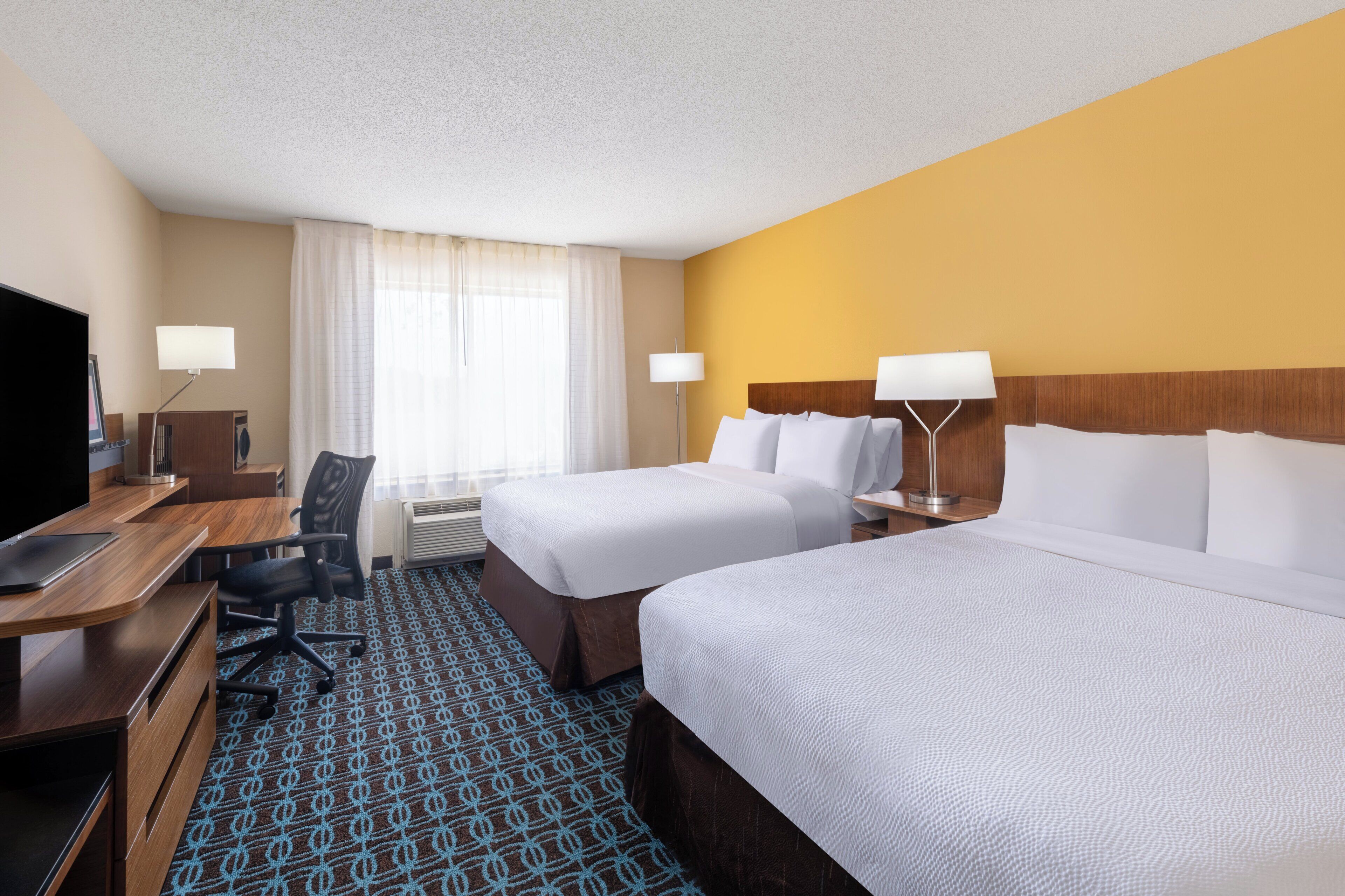 A hotel room with two beds, a desk with a chair, a television, and a window with curtains.