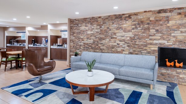 A modern lobby with a stone wall, a grey sofa, a brown leather chair, a round coffee table, and a TV mounted on the wall. - Garner Hotel Macon West by IHG (Macon)