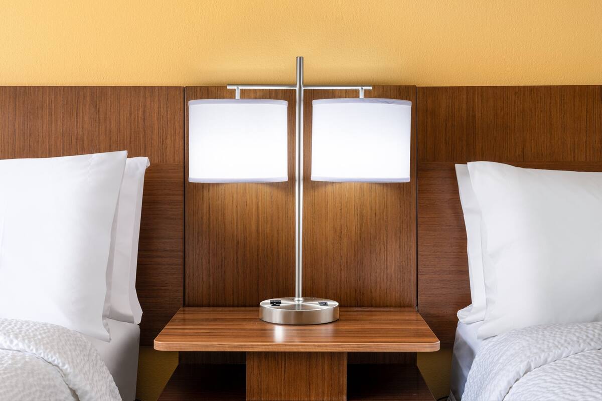 A hotel room with a wooden headboard, two beds with white linens, and a bedside table with a lamp.