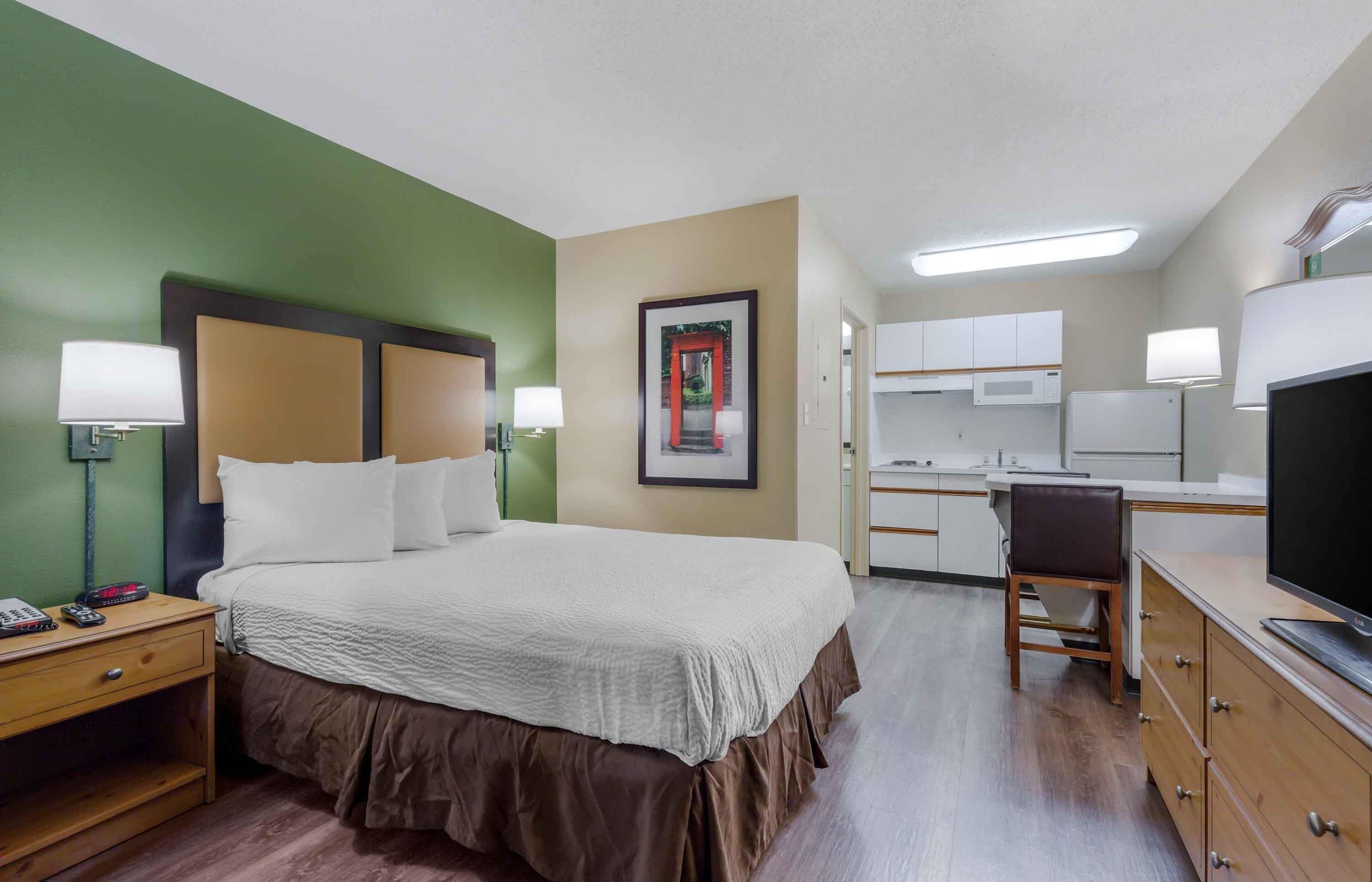 A hotel room with a bed, bedside tables, a desk, a chair, a television, and a kitchenette.