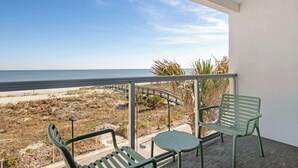 Varanda com duas cadeiras verdes e uma pequena mesa, com vista para uma praia e um calçadão.