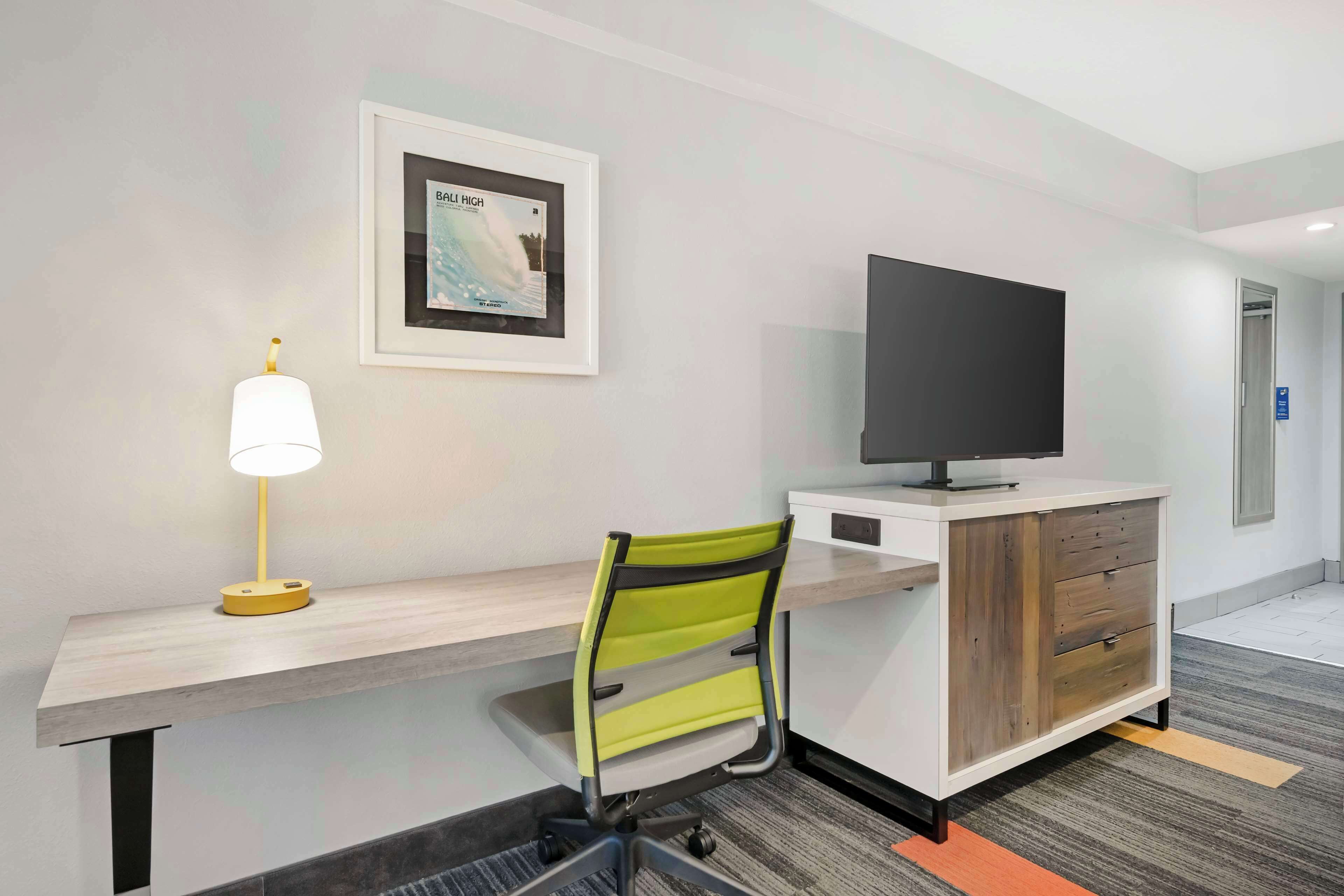A hotel room with a desk, a chair, a television on a cabinet, a framed picture on the wall, and a lamp.