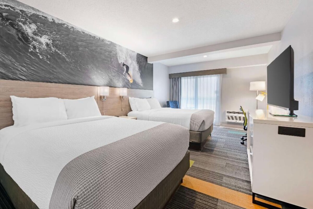Ein Hotelzimmer mit zwei Betten, einem großen Kunstwerk an der Wand, einem Schreibtisch mit Lampe und einem Fernseher.