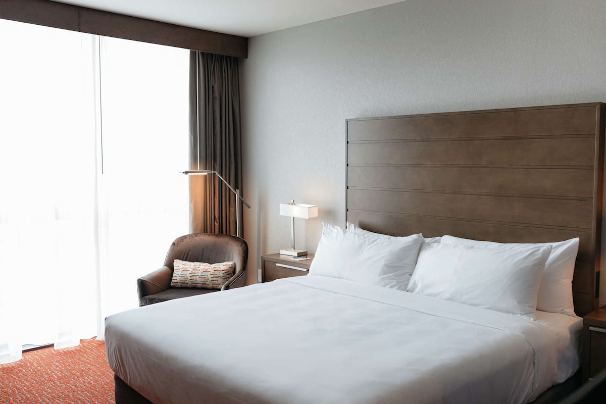 Ein Hotelzimmer mit einem großen Bett, einem Sessel, einem Nachttisch mit Lampe und Blick ins Freie.