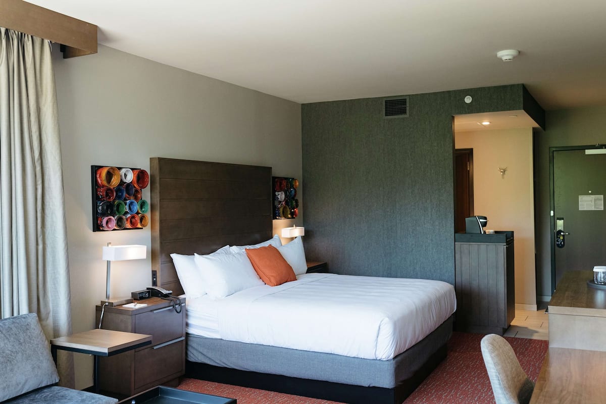 Ein Hotelzimmer mit einem großen Bett, Nachttischen, einem Schreibtisch und einem Stuhl. An der Wand hängt ein Gemälde und eine Tür führt in einen weiteren Raum.
